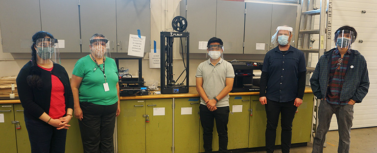 Photo of five people facing the camera, standing in front of three 3D printers. Each wears a face mask and a face shield