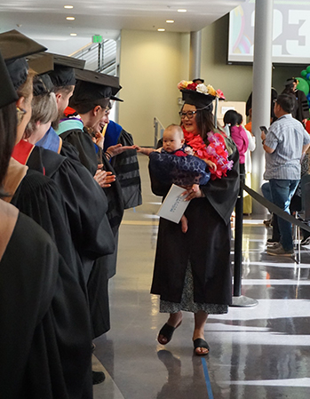 Highline College graduate holding baby walks down the receiving line at Commencement.